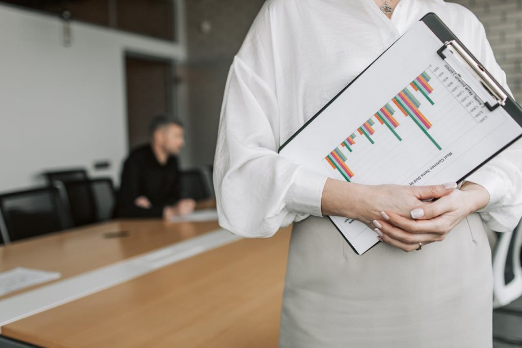 person-in-white-long-sleeve-shirt-holding-a-clipboard-with-statistics-7433922 Close-up of a person holding a clipboard with charts in a meeting room.